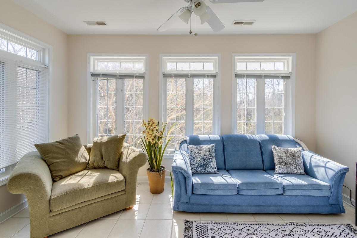 A light-filled four-season room features two sofas, one in soft beige and the other in blue. Large windows provide ample natural light, overlooking scenic views. A potted plant adds a touch of greenery, enhancing the room's inviting ambiance.