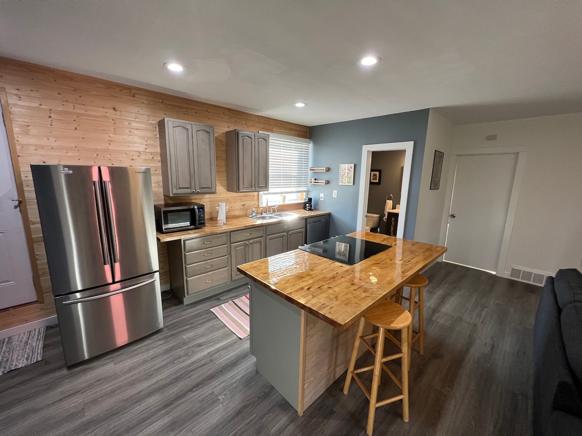 The kitchen area features a modern design with wooden cabinetry and a large island with seating for three. Stainless steel appliances, including a refrigerator and microwave, complement the space. Natural light enters through the window, illuminating the warm wood tones and neutral colors.
