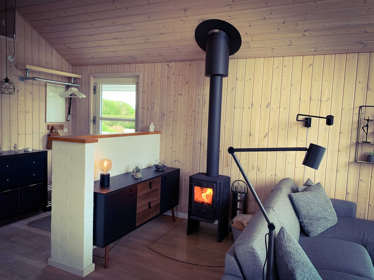 A cozy living area is highlighted by a contemporary wood stove, providing warmth. Light wood paneling contrasts with dark furnishings, including a sleek sofa and a stylish sideboard. A large window allows natural light and views of the surrounding nature, enhancing the inviting space.