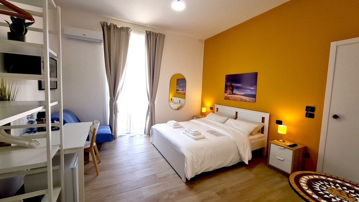 A bright room features a spacious bed with crisp white linens. One wall is painted in a warm yellow hue, while large windows are dressed with light curtains. A desk and chair provide a workspace, and a small seating area enhances the inviting ambiance.