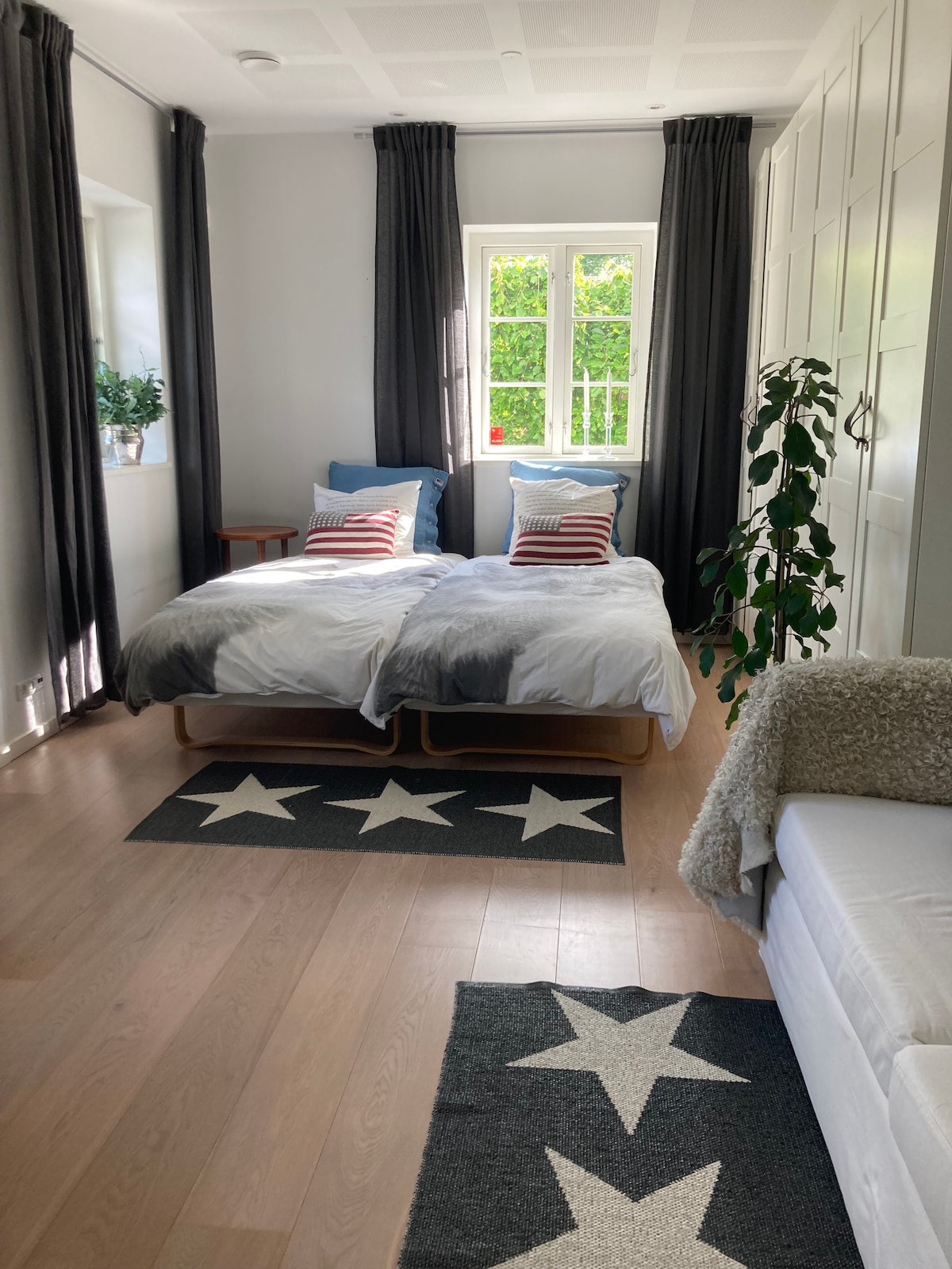 A bright bedroom features two spacious beds with light bedding, accented by striped pillows. A small wooden table sits beside the beds, while a green plant adds a natural touch. Large windows are dressed in dark curtains, allowing soft light to illuminate the room.