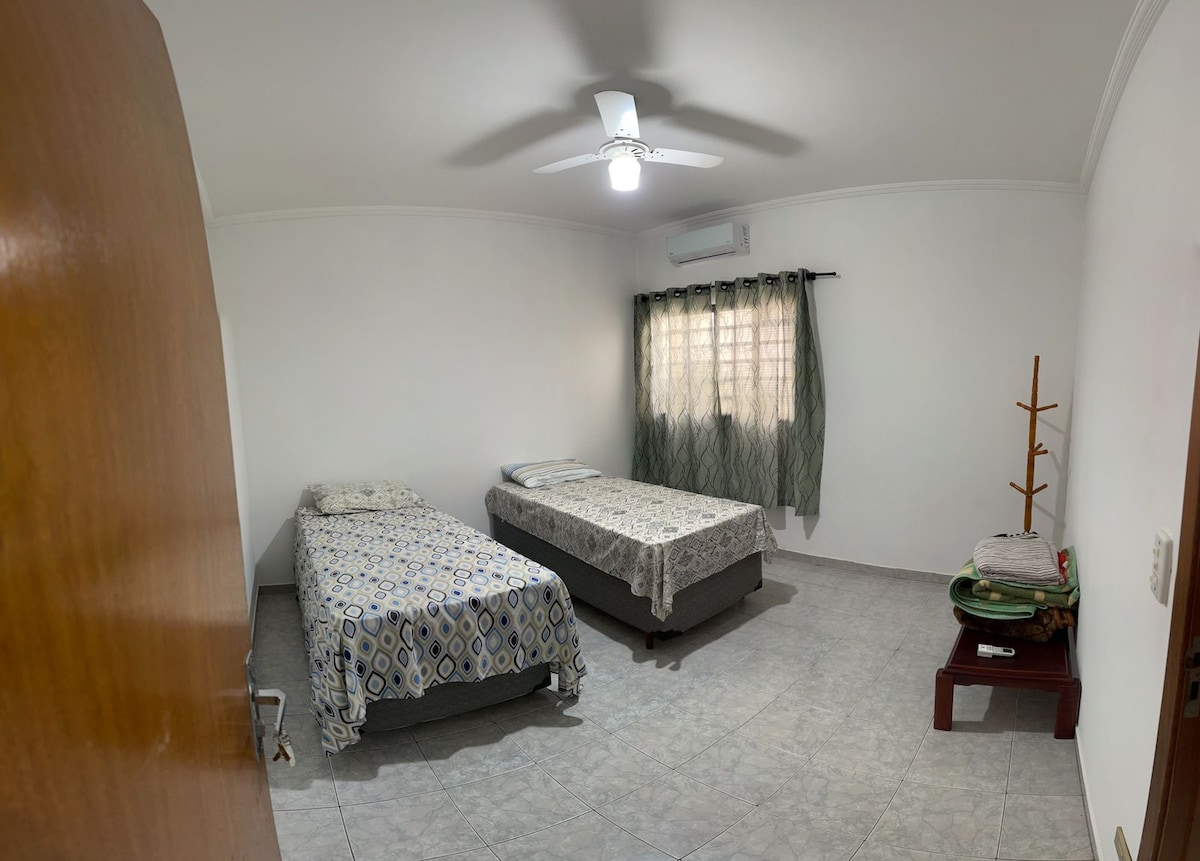 A well-lit bedroom features two comfortable twin beds, each with patterned bedding. A ceiling fan is visible above, providing airflow. Natural light filters through a window with sheer curtains. A simple wooden stand and a small table contribute to the room's functional layout.
