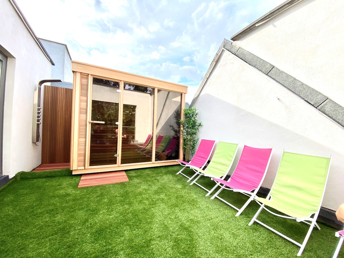 A serene outdoor space features a wooden sauna surrounded by green grass. Five lounge chairs in bright pink and green colors are arranged for relaxation. The sky above displays soft clouds, creating a calm atmosphere for unwinding.