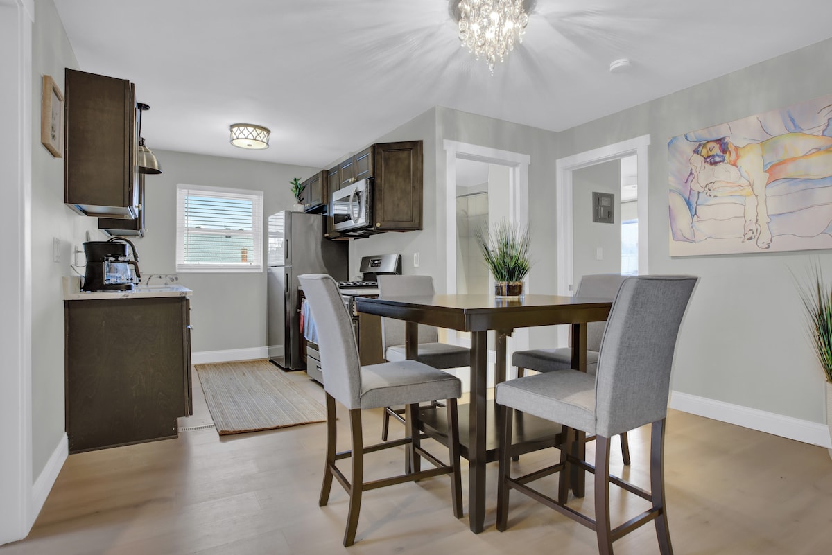 A dining area is presented with a sleek table and four upholstered chairs. The kitchen features modern appliances, including a refrigerator and an oven, complemented by a window allowing natural light to fill the space.