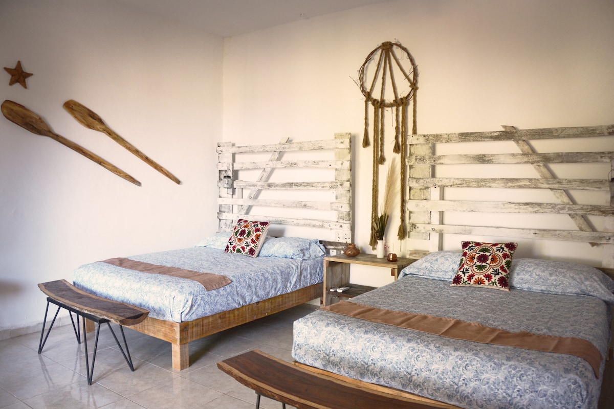 A spacious sleeping area displays two beds with decorative pillows. The headboards are crafted from rustic wood, while a wooden bench is positioned at the foot of each bed. Artistic elements, including dreamcatchers and wooden paddles, are mounted on the walls.