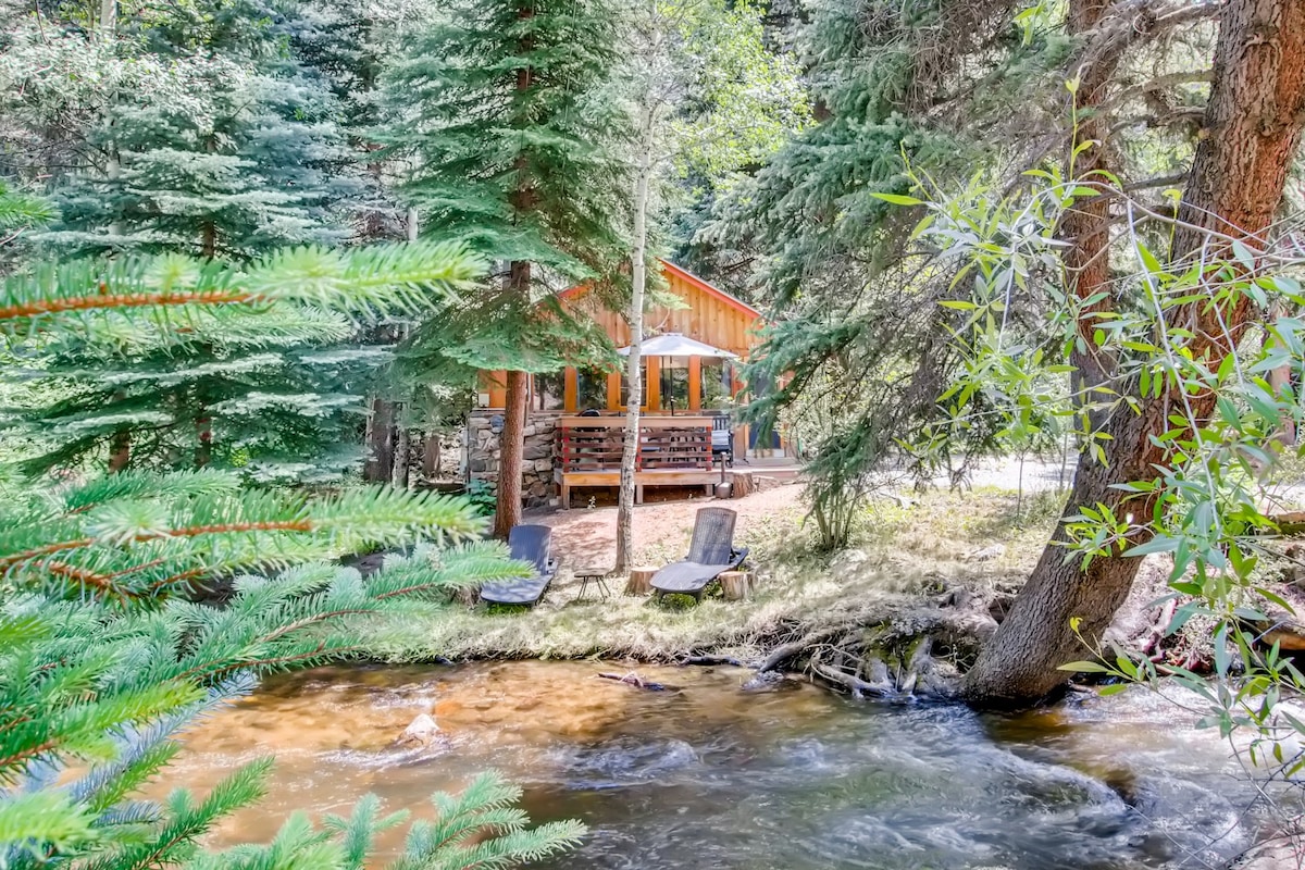 A rustic cabin, surrounded by lush green trees, is visible near a gently flowing creek. Adirondack chairs are positioned on the bank, providing a tranquil spot to enjoy the natural beauty. Sunlight filters through the foliage, casting dappled light on the water and surrounding area.