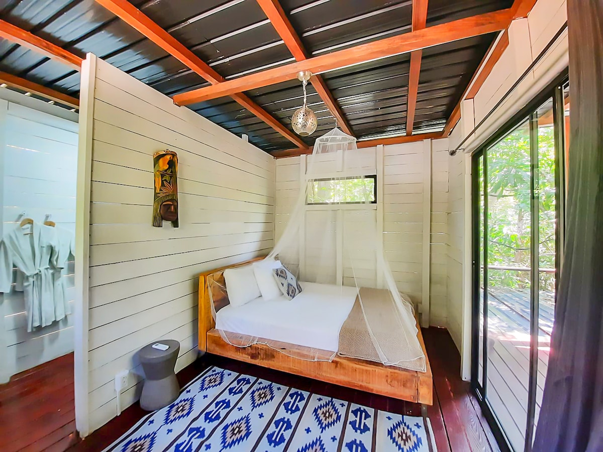 A cozy sleeping area is featured, showcasing a spacious bed with a decorative headboard and a light canopy. Natural light streams in through a large window, while a vibrant area rug adds a pop of color beneath the bed. Nearby, robes are neatly arranged for guest use.