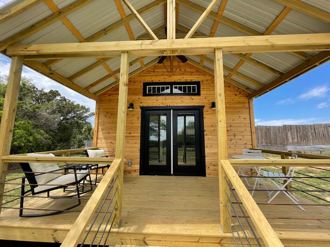 The cabin entrance features a spacious wooden deck with black rocking chairs and a small white table, framed by a decorative wooden railing. Double glass doors offer a welcoming entry, showcasing the cabin's natural wood exterior and high, vaulted ceiling.