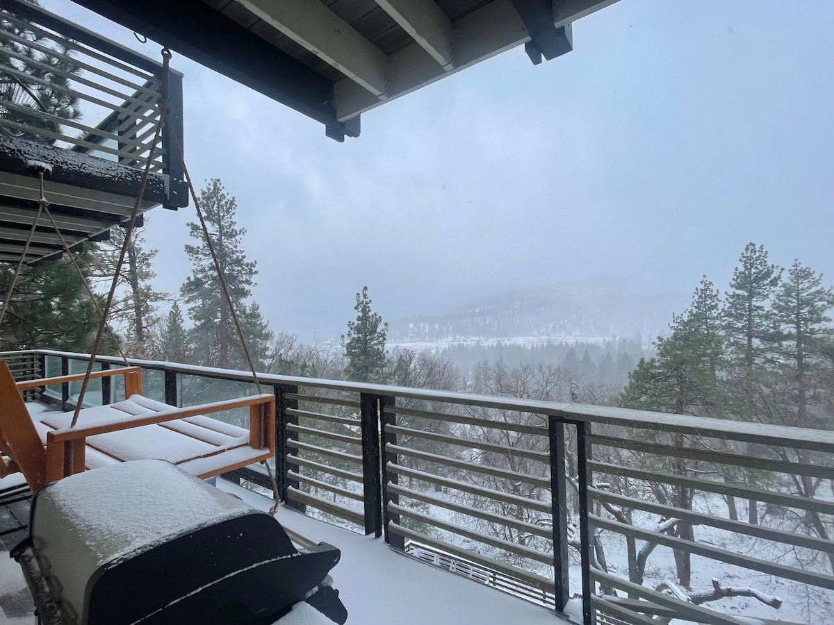 A covered outdoor deck features a swing and comfortable seating, both dusted with snow. The view includes a snowy landscape with pine trees, and distant mountains are barely visible through a mist. The atmosphere conveys a serene winter setting.