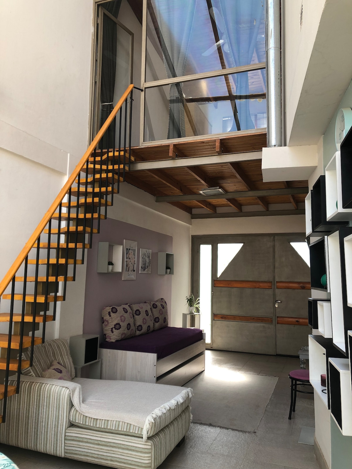 A spacious living area features tall ceilings with light streaming through large upper windows. A modern staircase with wooden steps leads to an upper level. A comfortable seating area includes a cozy diván and accent chairs, complemented by stylish wall art and a welcoming entrance door.