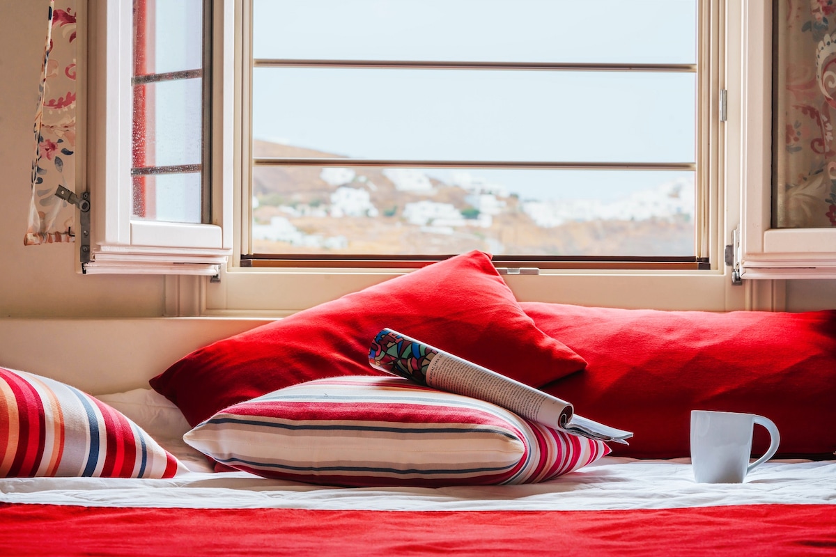 A cozy bed is layered with red and striped pillows, positioned near a window. Natural light enters through the open window, highlighting a rolled-up magazine and a white mug on the bed's surface. Soft curtains frame the window, offering views of the surrounding landscape.