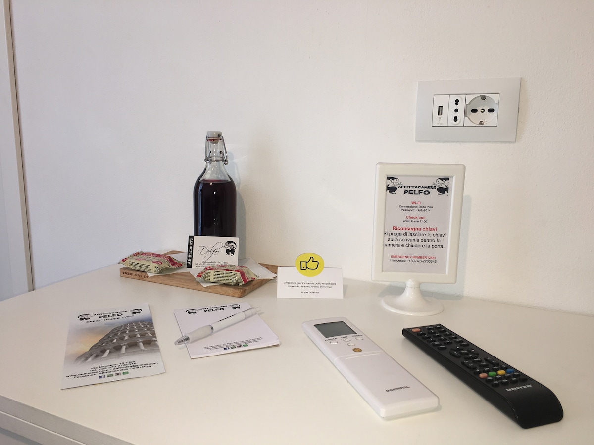 A neatly arranged countertop features a bottle of local beverage, a tray of snacks, a remote control, and informational materials about the property. A digital device stands ready for use, and a small sign provides guest instructions. The setting is bright and minimalistic.