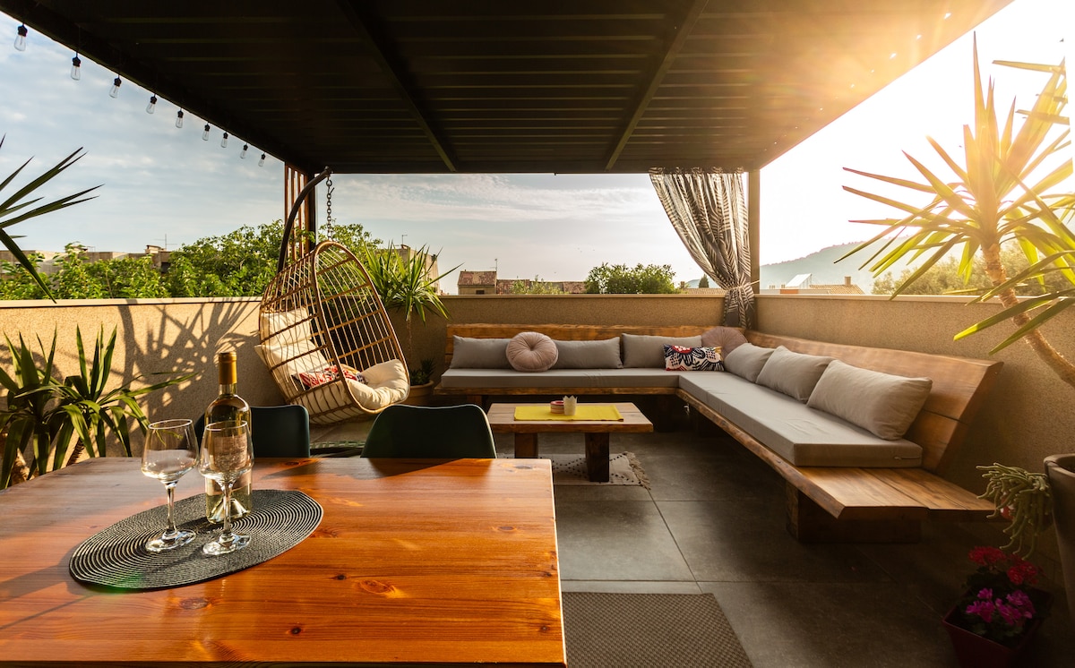 A spacious terrace is furnished with a large seating area and a wooden dining table. Potted plants add greenery, while warm sunlight casts a soft glow. A cozy hanging chair and soft cushions enhance the inviting atmosphere, providing an outdoor space for relaxation.