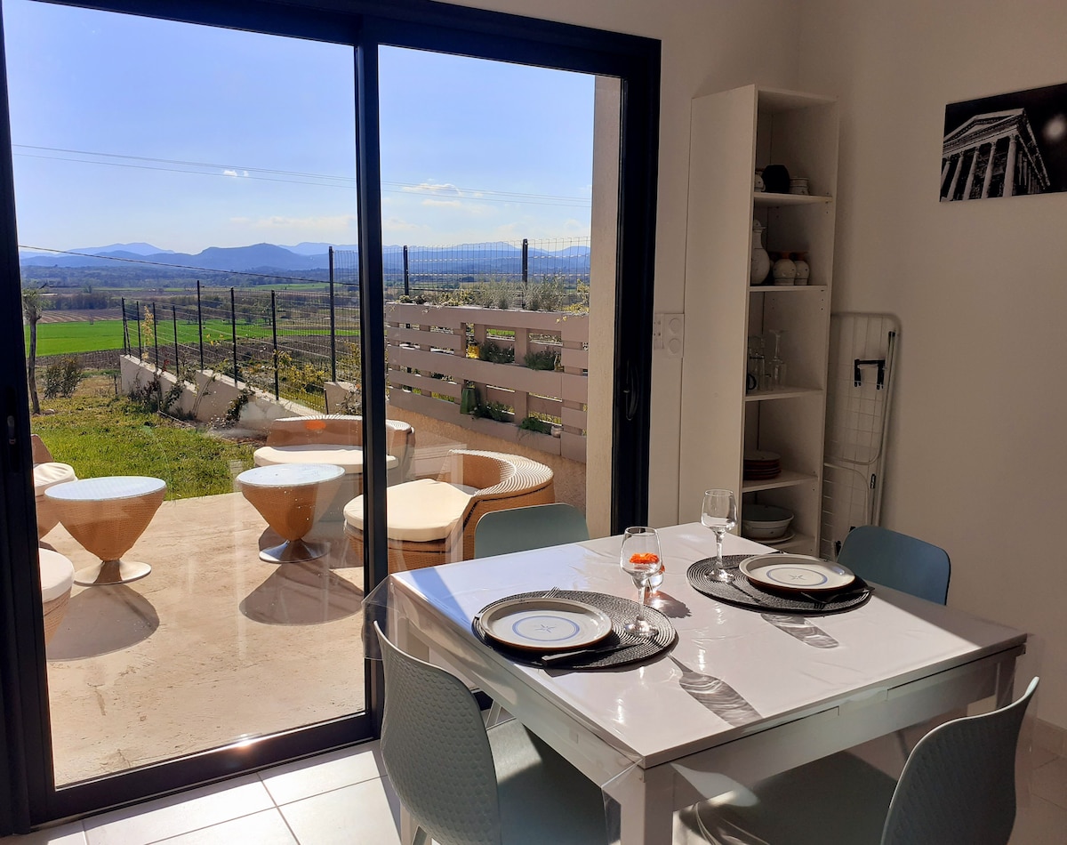 A dining area features a white table set for two, with plates and a glass placed on top. Large glass doors open to reveal a view of rolling green fields and distant mountains. Outdoor seating can be seen on the patio, enhancing the light-filled space.
