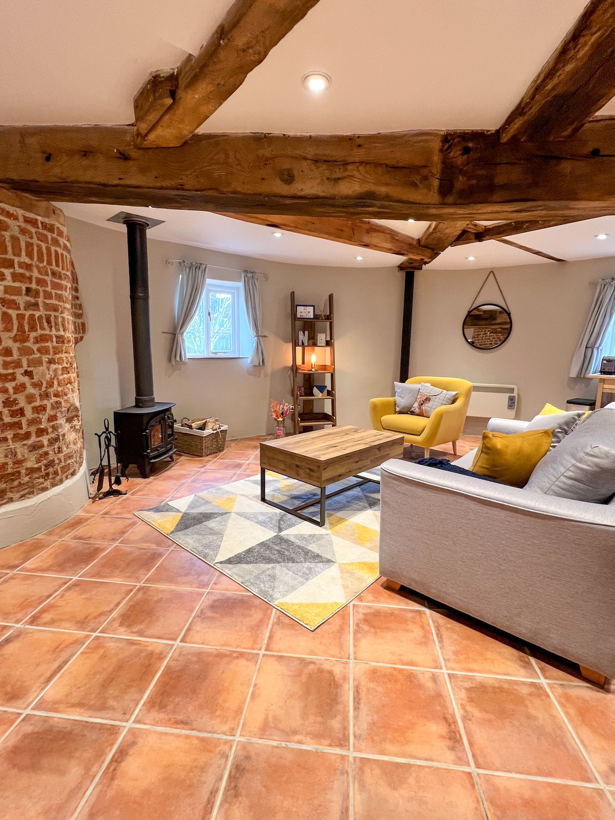 The open-plan living area features warm terracotta tiles and exposed wooden beams. A cozy sofa and armchair are arranged around a coffee table. A small wood-burning stove enhances warmth, while light enters through windows dressed with soft curtains.