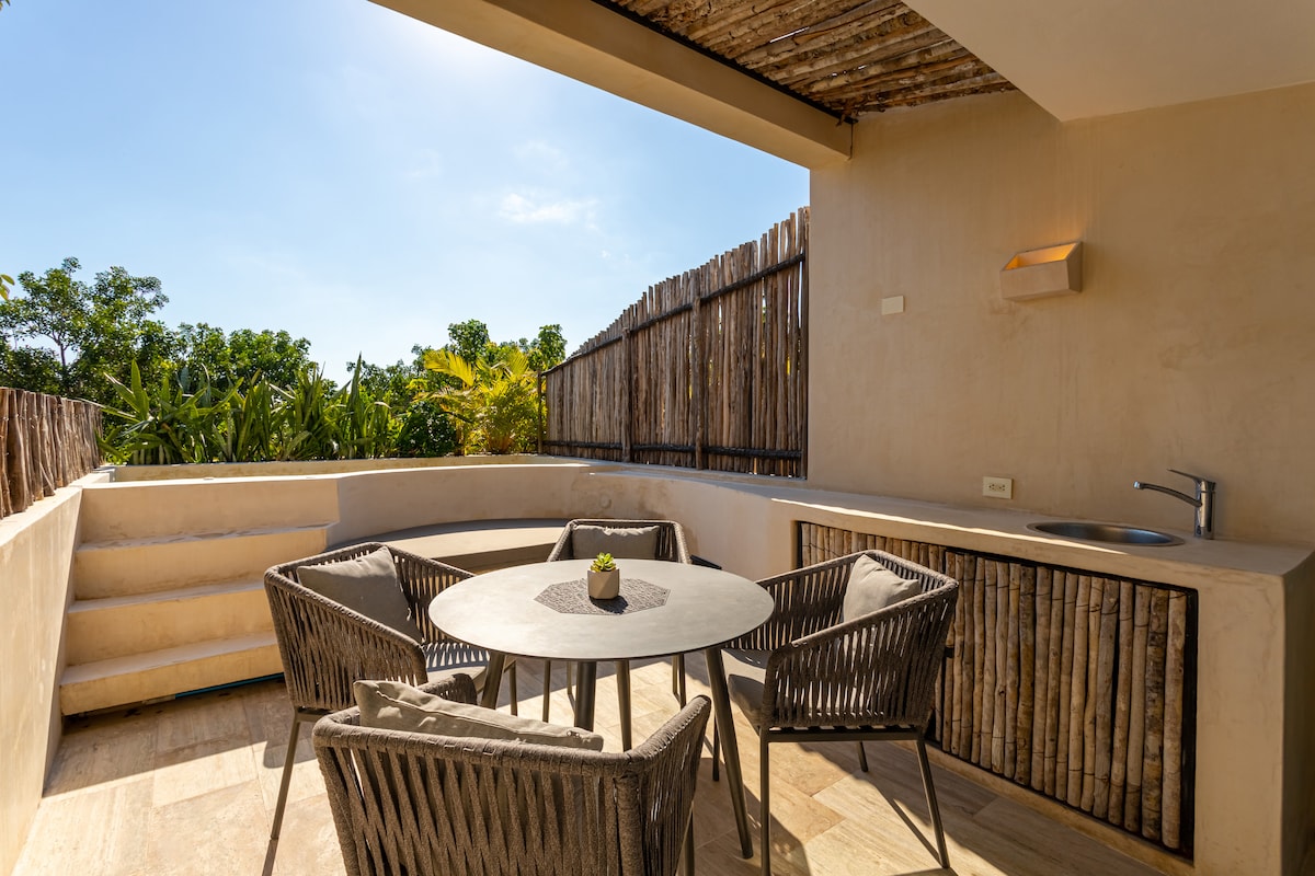A private terrace features a circular table surrounded by four woven chairs. A small sink and counter space are situated nearby, with steps leading down to the lower level. Lush greenery can be seen in the background under a clear blue sky.
