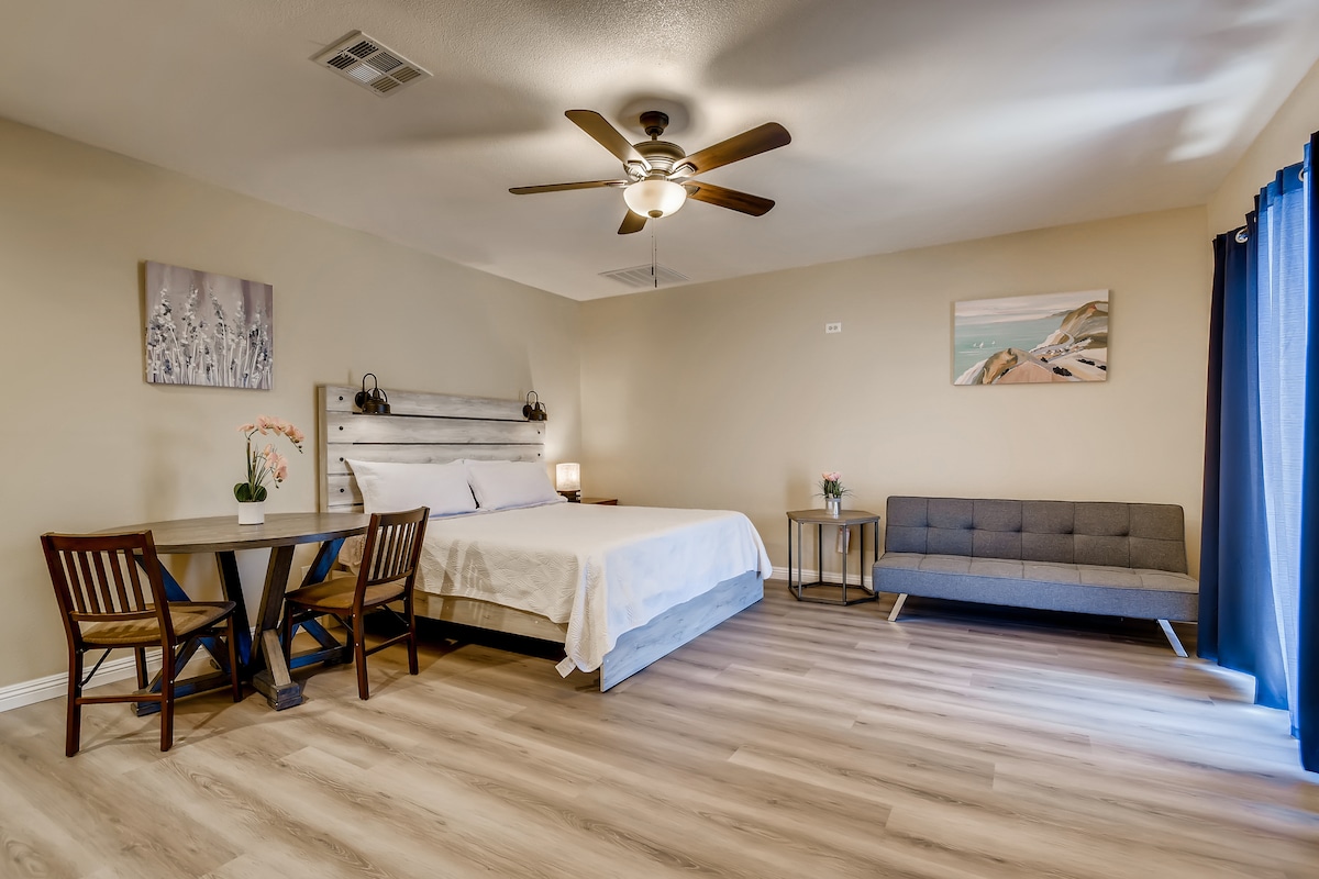 A spacious studio features a king bed near a table with two chairs. A comfortable sofa is positioned against the wall. A ceiling fan is installed above, and natural light enters through the curtain-covered window, enhancing the light wood-colored floor.