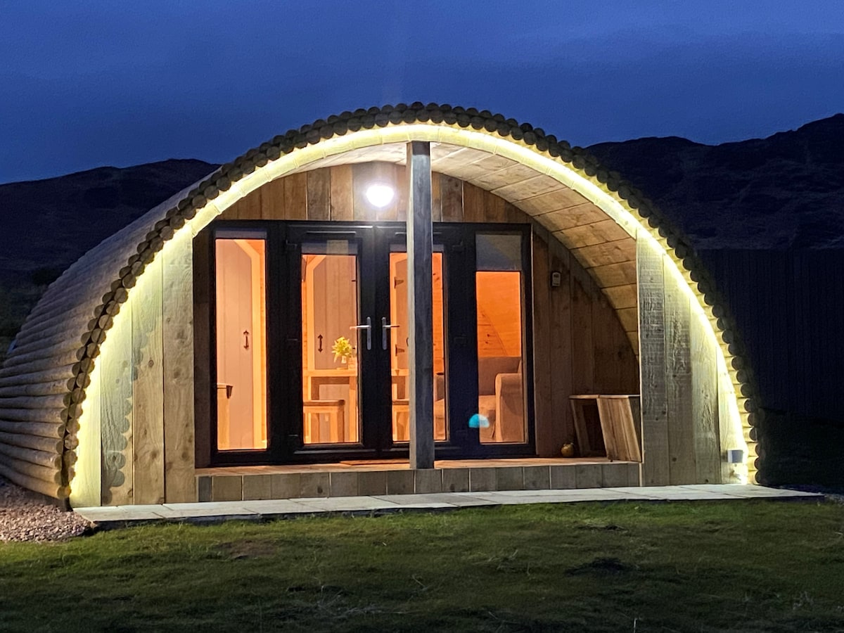 A modern cabin is illuminated by soft outdoor lighting, showcasing a curved wooden structure. The front features large glass doors leading to a warm interior glow, surrounded by a grassy area. The backdrop includes rolling hills, enhancing the cabin's natural setting.