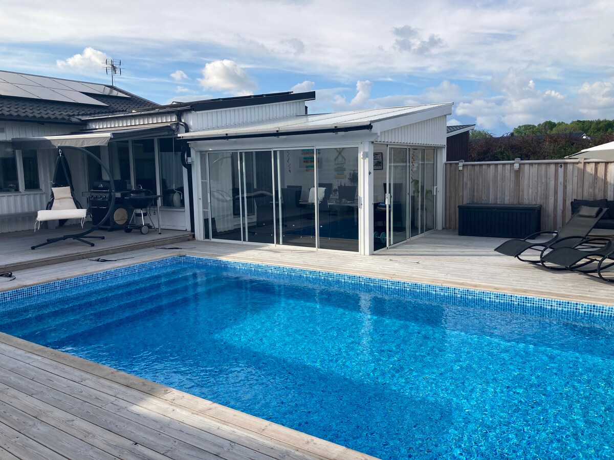 A spacious outdoor area features a clear blue pool surrounded by a wooden deck. Adjacent, a glass-enclosed sunroom provides a seamless connection to the main living space. Comfortable lounge chairs are positioned for relaxation under the sun, with a grill visible in the background.