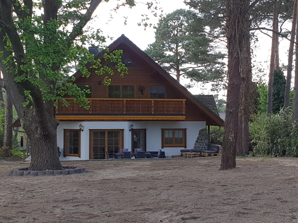A modern wooden house is framed by tall trees, featuring a spacious balcony above the ground floor. Seating areas are positioned on the sandy yard in front of the house, creating an open and inviting space. The roofline is distinctly angled, contributing to its unique design.