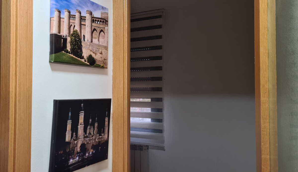 Two framed photographs are displayed on the wall. One shows a castle-like structure, while the other captures the illuminated towers of a historical building at night. A window with light-filtering blinds is visible, casting gentle shadows in the room.