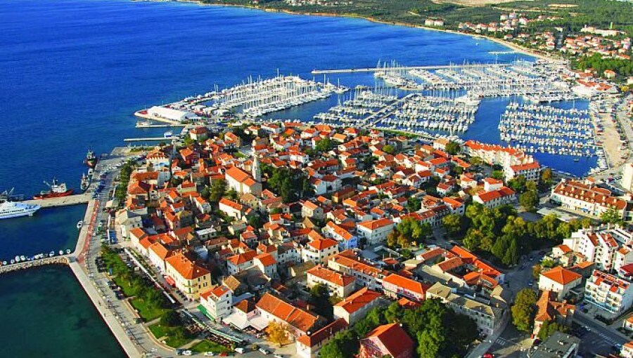 An aerial view captures a coastal town with terracotta rooftops and a marina filled with numerous boats. The vibrant blue waters contrast with the lush greenery and surrounding hills, while the spacious harbor is visible on the right, enhancing the scenic beauty of the area.
