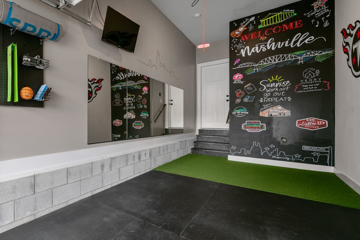 An entryway is featured, showcasing a private gym area with green turf flooring and black rubber mats. A large mirror reflects the space, while a television is mounted on the wall. Colorful wall art highlights Nashville's culture, creating an inviting atmosphere.