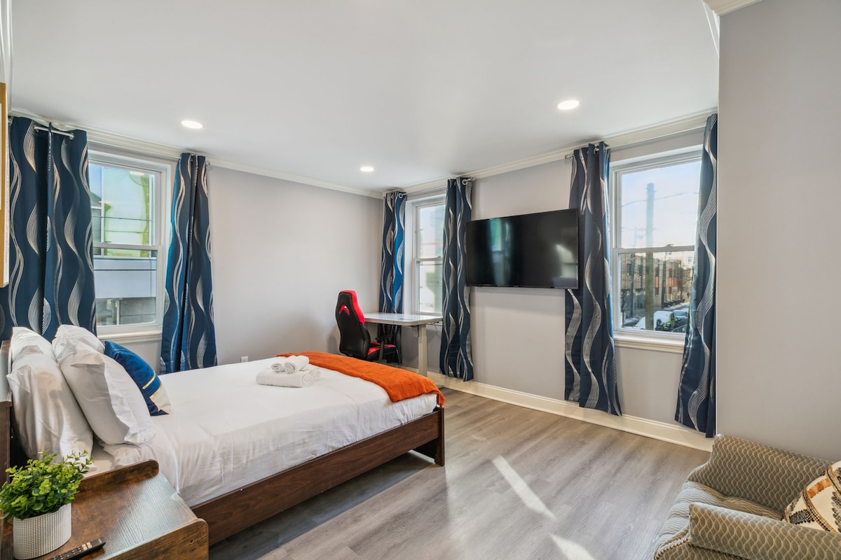 A bright bedroom features a spacious bed covered with crisp white linens and an accent throw. Large windows allow natural light to fill the room. A smart TV is mounted on the wall, and a small desk is positioned nearby, offering a functional workspace.