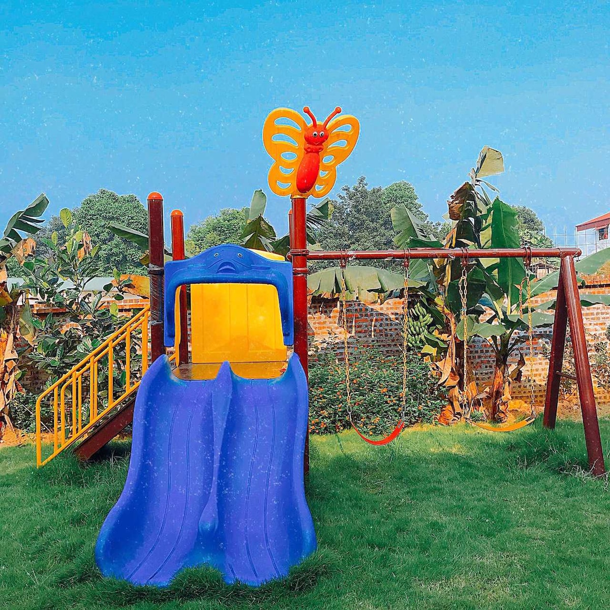 A colorful playground is featured, equipped with a vibrant slide and a swing set. The slide is shaped like a butterfly, set against a backdrop of greenery, including banana plants. The grassy area provides ample space for outdoor play.