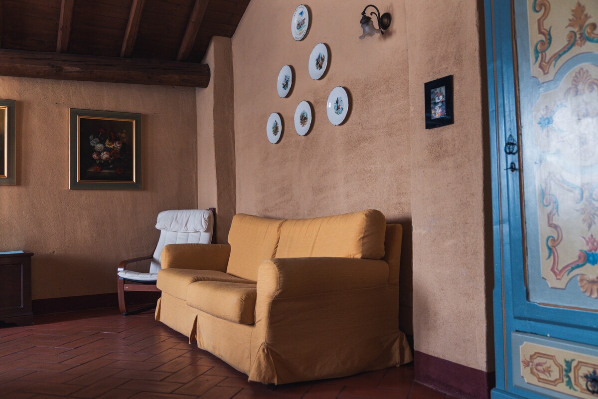 A cozy seating area is presented with a yellow couch and a white armchair. Decorative plates are arranged on the rustic wall, while artwork is displayed in a small frame. Exposed wooden beams and terracotta flooring add to the warm ambiance.