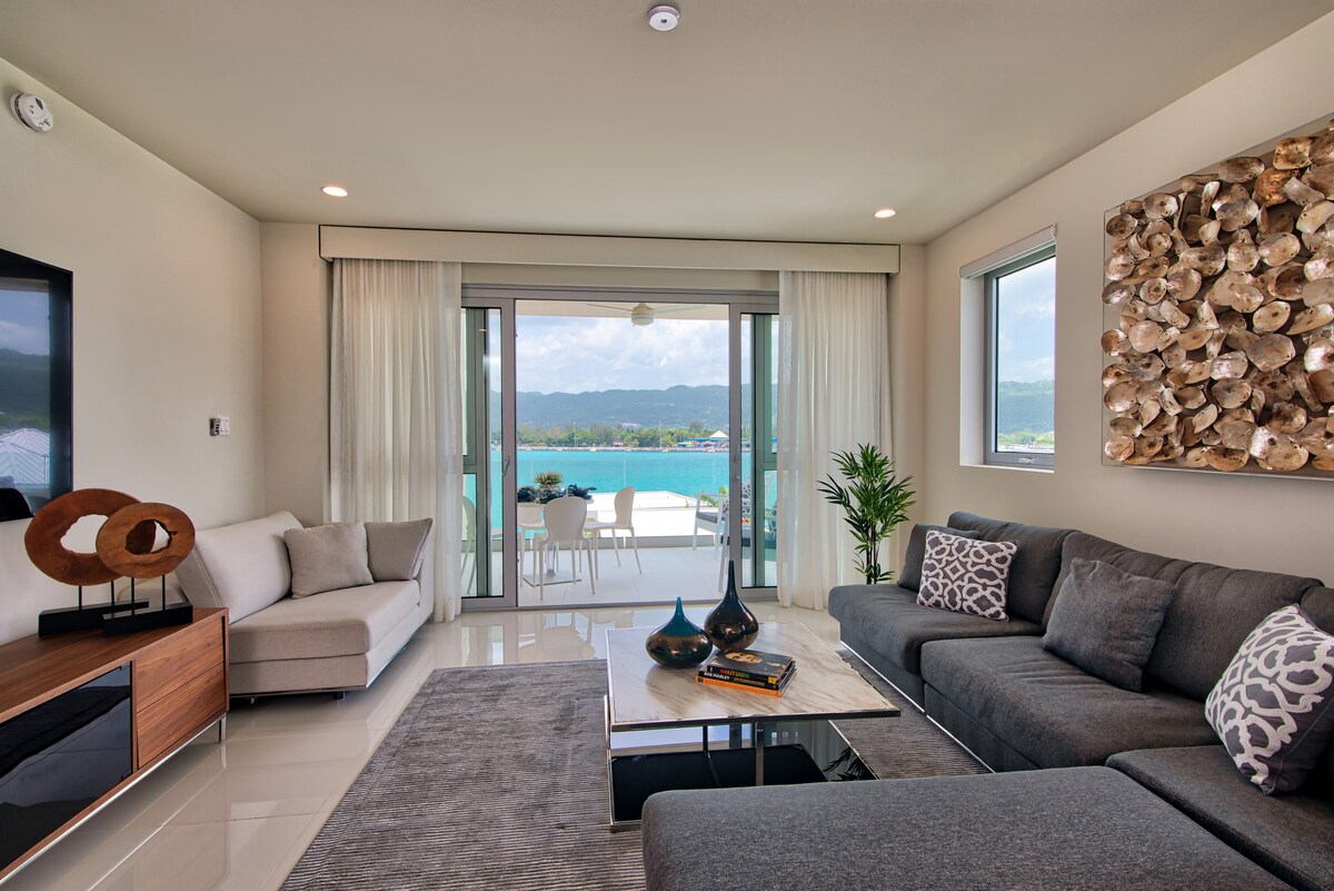 A modern living area is seen, featuring a large grey sectional sofa positioned around a sleek coffee table. Natural light pours in through the expansive glass doors, which open to stunning sea views. A decorative piece made of shells graces one wall, adding a coastal touch.