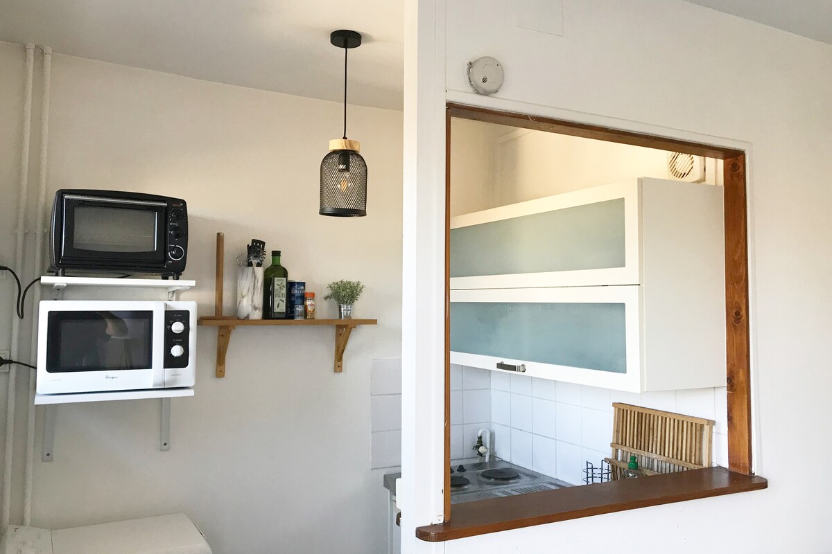 A compact kitchen area is presented, featuring a white microwave and a small black microwave above a wooden shelf. The kitchen is equipped with modern cabinets, a sink, and a small stove. Natural light illuminates the space through a window with minimalistic design.