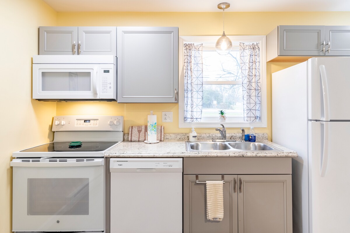 The kitchen features modern appliances, including a refrigerator, dishwasher, microwave, and stove. A window provides natural light, and cabinets in grey and white offer ample storage space. Countertops are finished in a light-colored stone, creating a functional cooking area.