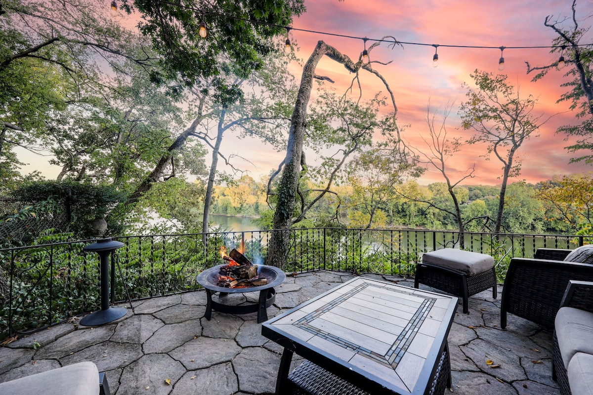 A stone patio offers a fire pit at its center, surrounded by comfortable seating. Twinkling lights hang above, set against a backdrop of vibrant sunset colors and lush greenery bordering the Cumberland River.