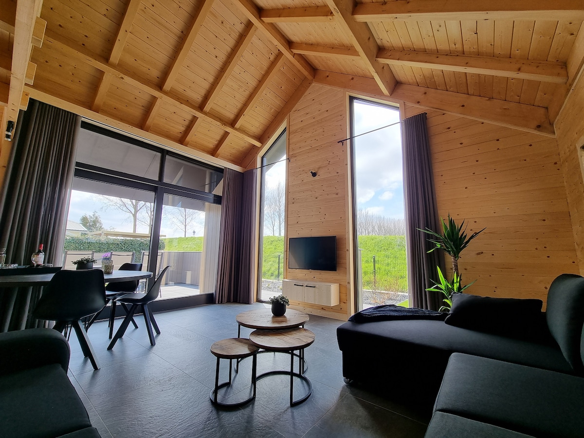 An open living area is shown with wooden walls and high ceilings. Large windows allow natural light to fill the space, revealing views of greenery outside. A dark sofa and small coffee tables are arranged comfortably, while a dining area is visible in the background.