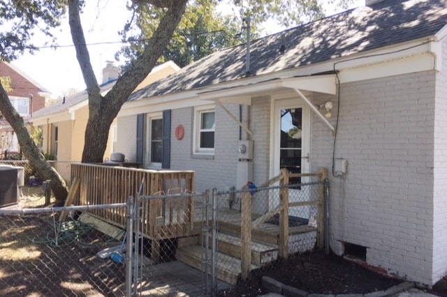 A single-story home with a light gray exterior features a wooden deck leading to a fenced entrance. The front door is complemented by a simple awning. Nearby, a large tree provides natural shade, enhancing the home's outdoor area.