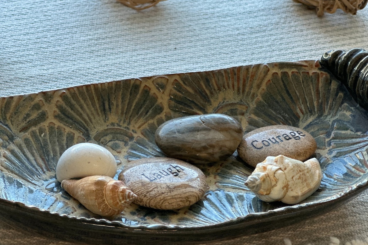 A decorative dish holds several smooth stones and shells arranged artistically. Two stones feature words etched into their surfaces, reading 'Laugh' and 'Courage.' The dish is set against a textured surface, adding to its aesthetic appeal.