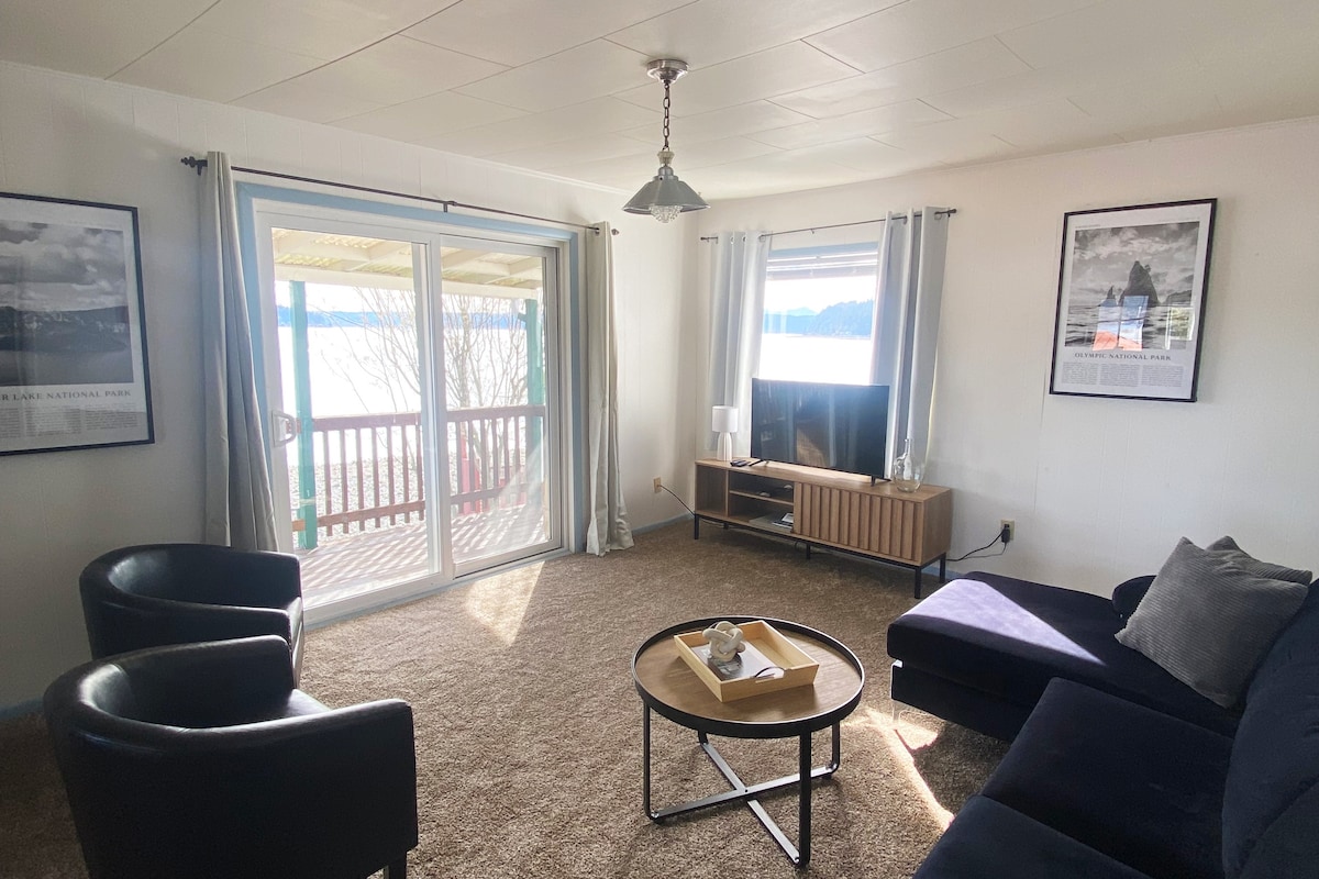 A cozy living area features a dark blue sofa and two black armchairs arranged around a circular table. Natural light enters through large sliding glass doors, leading to a deck. A television is positioned on a low wooden console, accompanied by framed art on the walls.