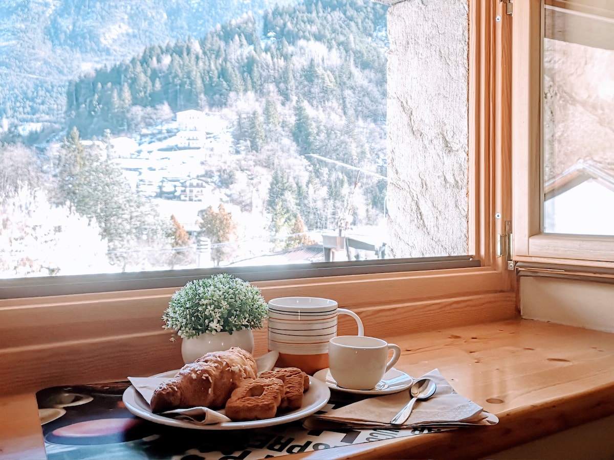 A wooden dining table is positioned near a window, offering a view of the mountainous landscape. A small potted plant adorns the table alongside a plate of pastries and a cup with a saucer. Natural light enhances the inviting atmosphere.