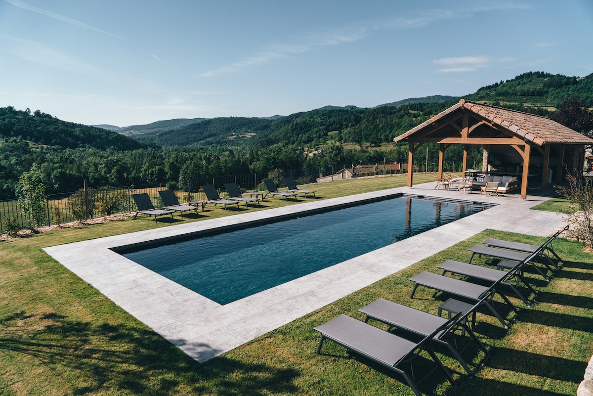 The outdoor area features a private, heated swimming pool surrounded by smooth stone decking. Six lounge chairs are arranged neatly along the poolside, facing expansive views of lush greenery and rolling hills. A covered pool house with comfortable seating is visible in the background.