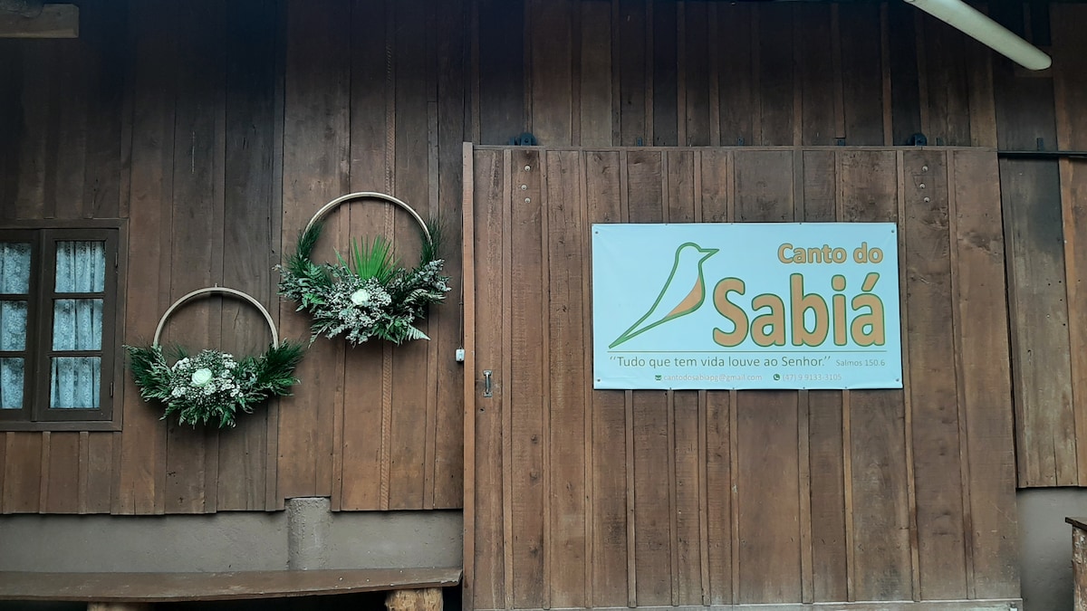 A rustic wooden exterior features two circular floral arrangements on either side of the entrance. A sign with the name 'Canto do Sabiá' hangs prominently, accompanied by a quote. The natural wood grain complements the greenery of the arrangements.