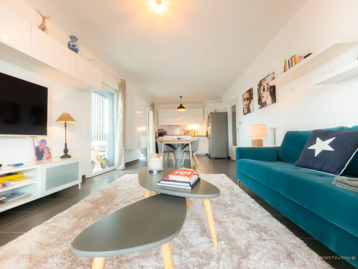 A contemporary living space features a cozy blue sofa and two nested coffee tables placed over a soft area rug. A modern television is mounted on the wall, and a lamp provides ambient lighting. The open kitchen is visible in the background, enhancing the communal feel.