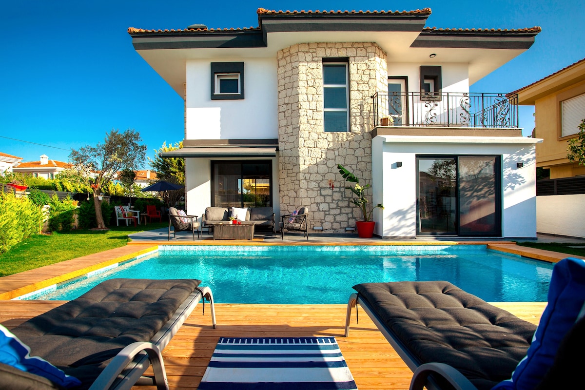 A two-story villa is presented with a stone exterior and large windows, overlooking a private pool surrounded by a wooden deck. The pool area features lounging chairs, and a lush garden is visible in the background, providing a serene outdoor space.