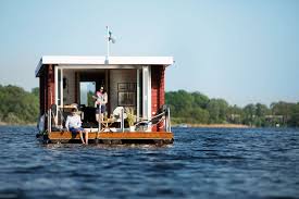 A floating house is seen on calm waters, featuring a wooden deck with a few chairs. The structure has large windows, allowing natural light to fill the interior. Surrounding greenery is visible, enhancing the natural setting.