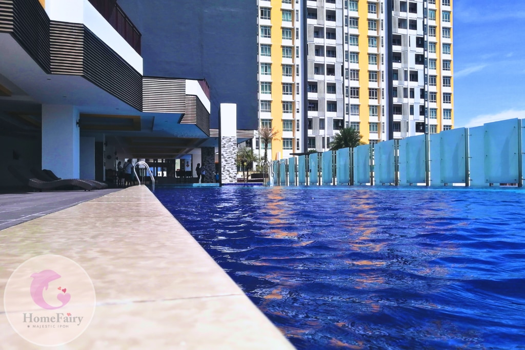 An inviting swimming pool is featured, with clear blue water reflecting sunlight. The poolside is lined with lounge chairs, and glass panels provide a sense of openness. Towering residential buildings are visible in the background, enhancing the atmosphere of a vibrant community.