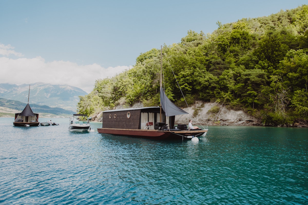 Two floating cabins are anchored on calm blue waters, surrounded by lush green trees. A small boat is visible, navigating the serene lake, while gentle waves reflect the sunlight. The picturesque backdrop features distant mountains, creating a peaceful aquatic setting.
