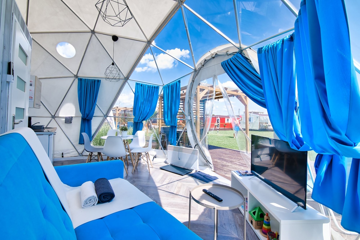 An inviting dome interior features a bright blue sofa and a dining area with a round table. Large windows allow natural light to fill the space, with vibrant blue curtains offering a pop of color. A flat-screen TV is positioned on a small white table.