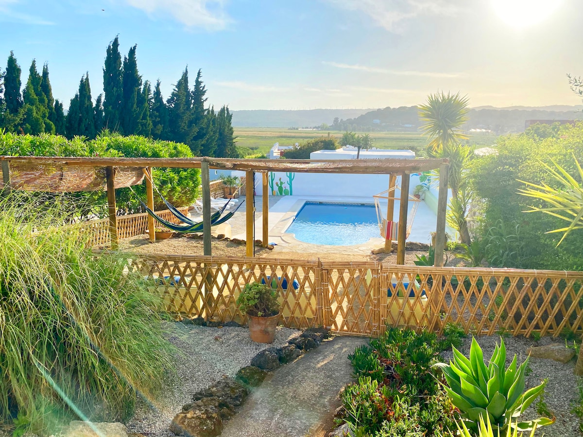 A serene outdoor area features a swimming pool surrounded by wooden decking and lush greenery. Two hammocks are visible, providing a relaxing spot under the sun. A wooden fence and various plants frame the view, enhancing the natural setting and inviting tranquility.
