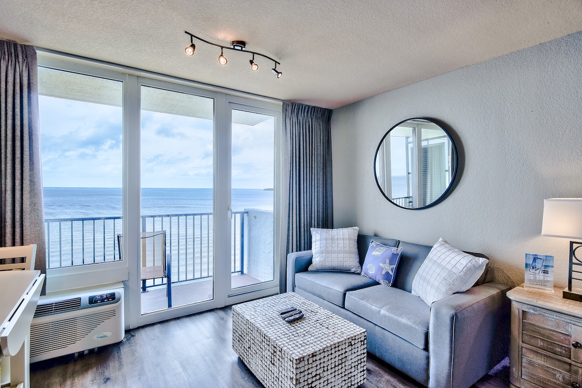 A bright living area features a comfortable gray sofa with decorative cushions, a round mirror, and a stylish coffee table. Floor-to-ceiling glass doors provide a stunning view of the bay, leading to a private balcony. Soft lighting complements the relaxed coastal ambiance.