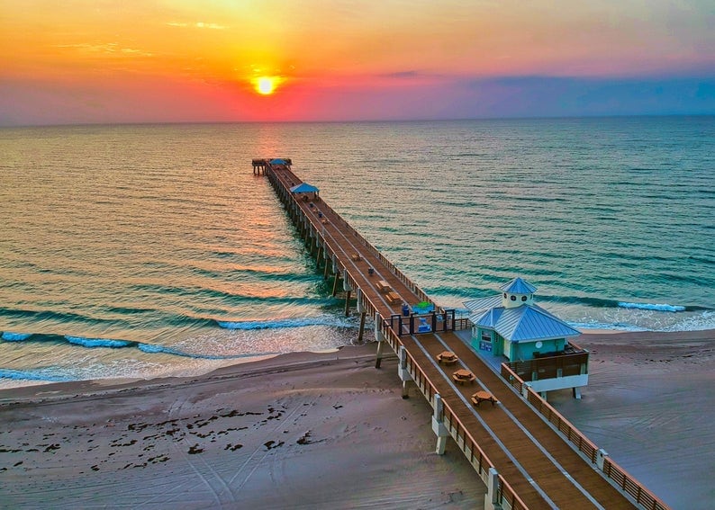 A wooden pier extends over calm waters, leading towards the horizon where a vibrant sunset casts hues of orange and pink across the sky. The gentle waves lap at the shoreline, with a small pavilion visible at the pier's end.